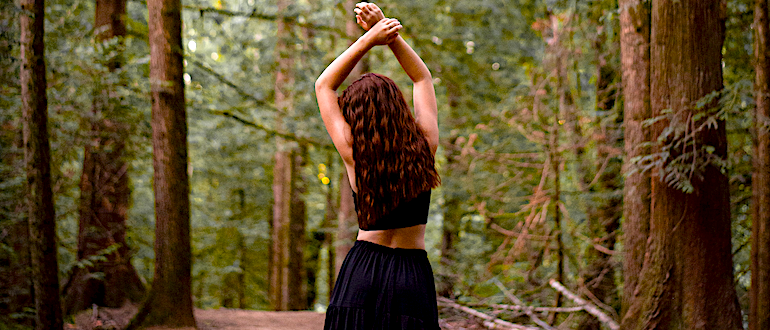 Woman with her back to you sniffs her left armpit, with a thick forest in the background